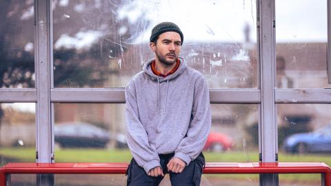 A man in a grey hoody and woolen hat sits at a bus stop looking thoughftul.