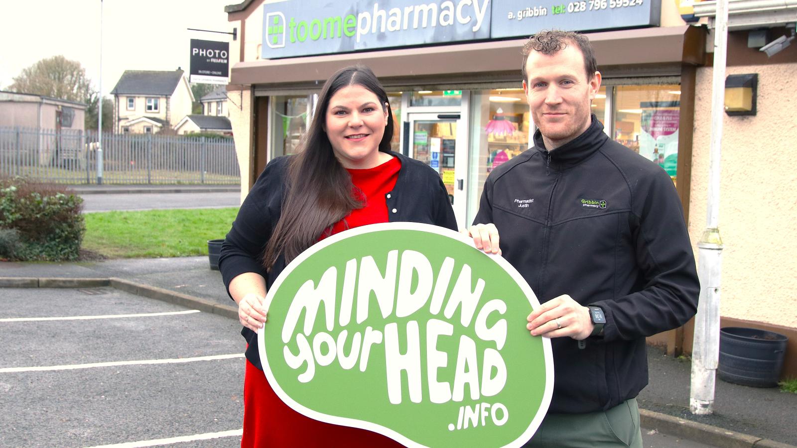 The Public Health Agency's Kathy Owens with pharmacist Justin Crozier standing outside Toome Pharmacy holding a green sign that says "MindingYourHead.info"